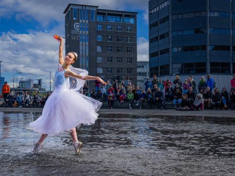 Ballerína dansar í vatni fyrir utan Hörpu. Seðlabankinn og mannfjöldi í baksýn, stúlkan er í hvítum ballettbol og tjullpilsi.