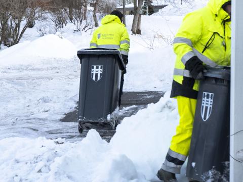 Two waste management employees pull bins marked with the city's logo, with snow outside.