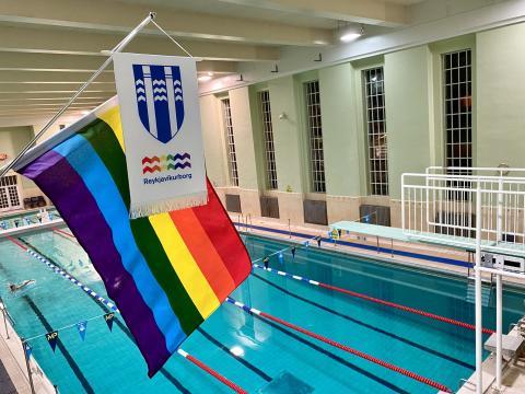 Interior of Sundhöll Reykjavíkur. Image shows the large pool with the high diving board on the far right. Prominently displayed in the foreground are the city flag and a larger flag, the rainbow flag (old version)