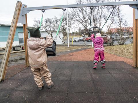 Two preschool children playing on swings on preschool grounds. They are wearing rain overalls, holding separate swings and facing away from the camera.
