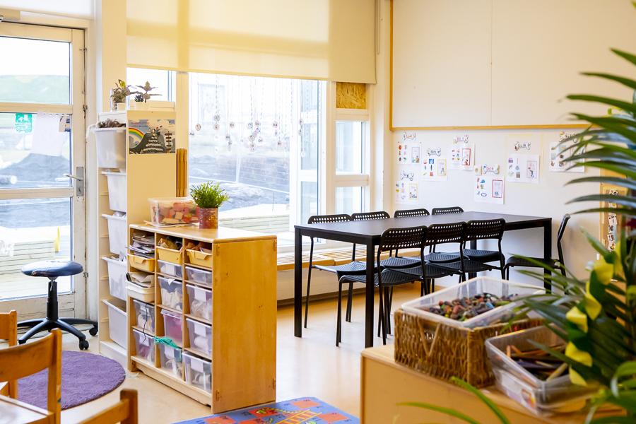 Séð inn í eitt af herbergjunum á leikskólanum Klömbrum. Svart borð og stólar, hillur með leikföngum og fleiri húsgöng.
