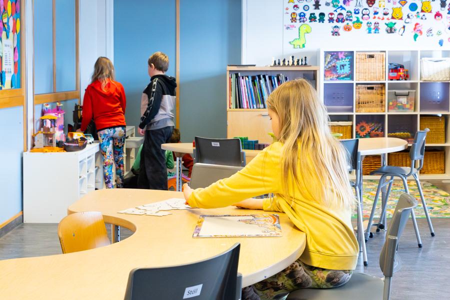 Barn situr við borð að pússla. Tvö börn standa við hillu að leika sér með dúkkuhús. Hillur með leikföngum í baksýn.