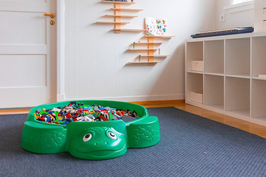 Stór plastskjaldbaka, á gólfi, sem fyllt er með legó kubbum. Hillur til hliðar og grá motta á gólfi. 