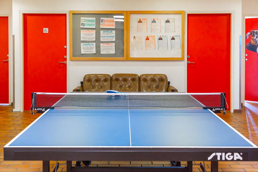 Salur með borðtennisborði, sófa og tilkynningum á vegg. Rauðar hurðir í baksýn. 