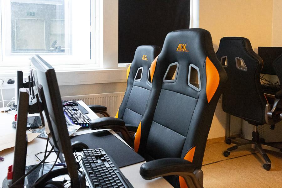 Room with computers on desks and black and orange office chairs.