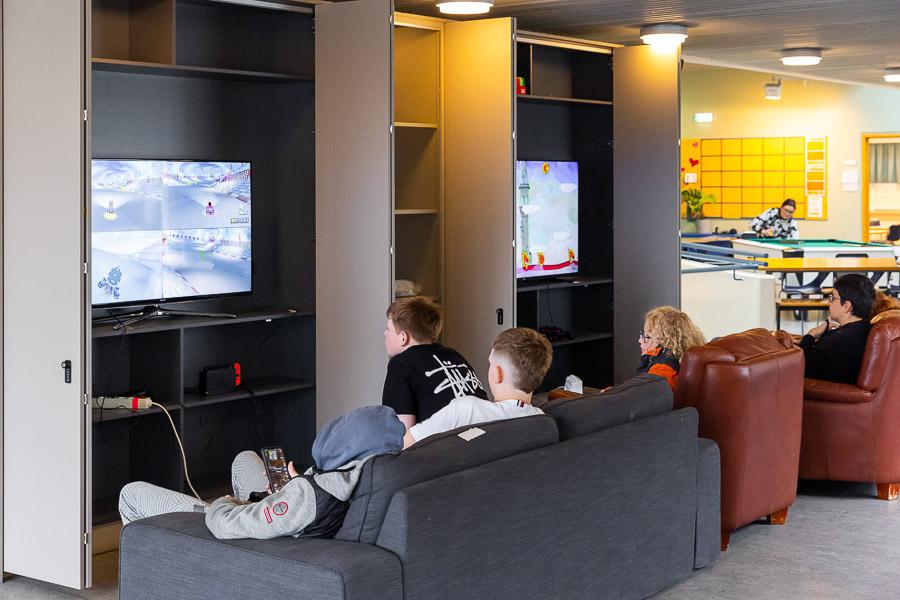 Children sitting on sofas in front of wall-mounted screens.