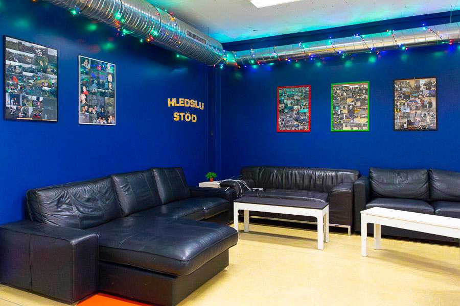 Corner with blue walls and a black corner sofa. The wall reads “Hleðslustöð” or charging station. Pictures hang on the wall nearby.