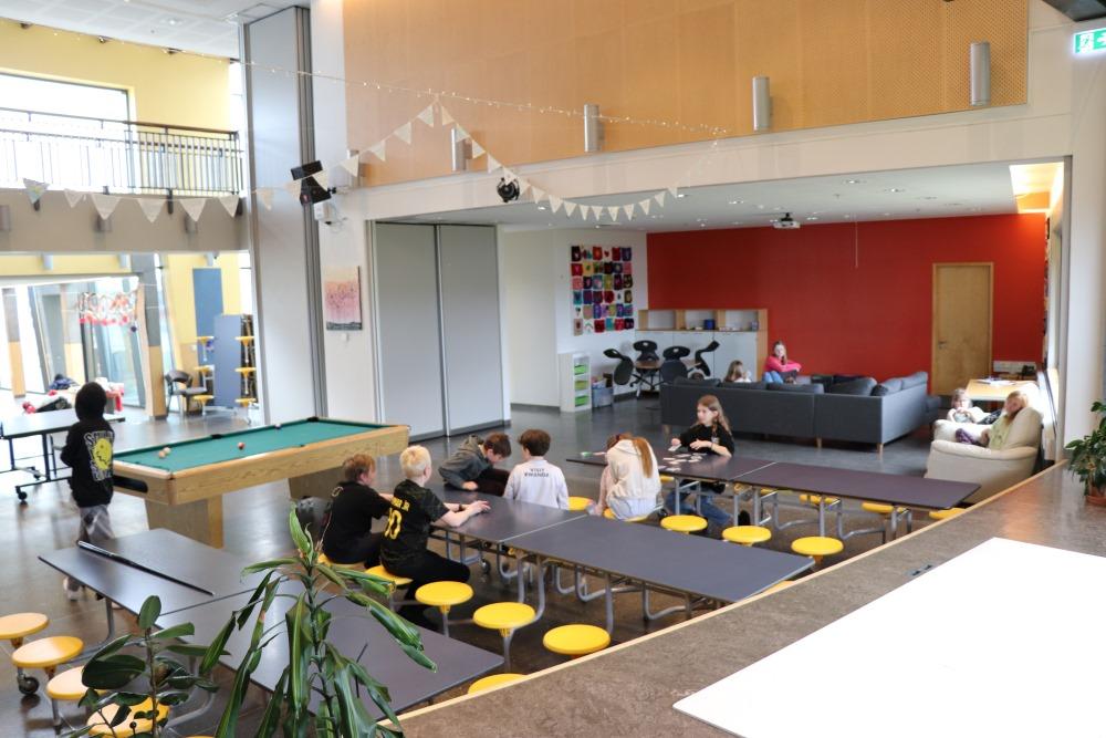 A large room with long tables, yellow stools, a pool table, couches and flags hanging from the ceiling. 