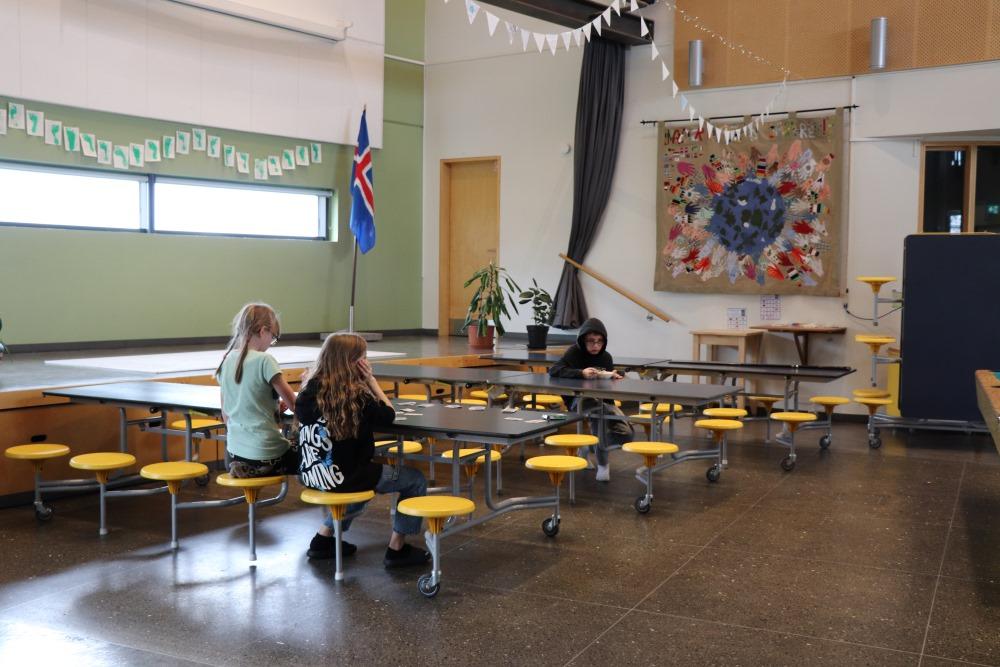 Long tables arranged in rows with yellow stools. Children playing and eating at the tables. Flags hang from the ceiling, with the Icelandic flag on the side and a picture on the wall. 