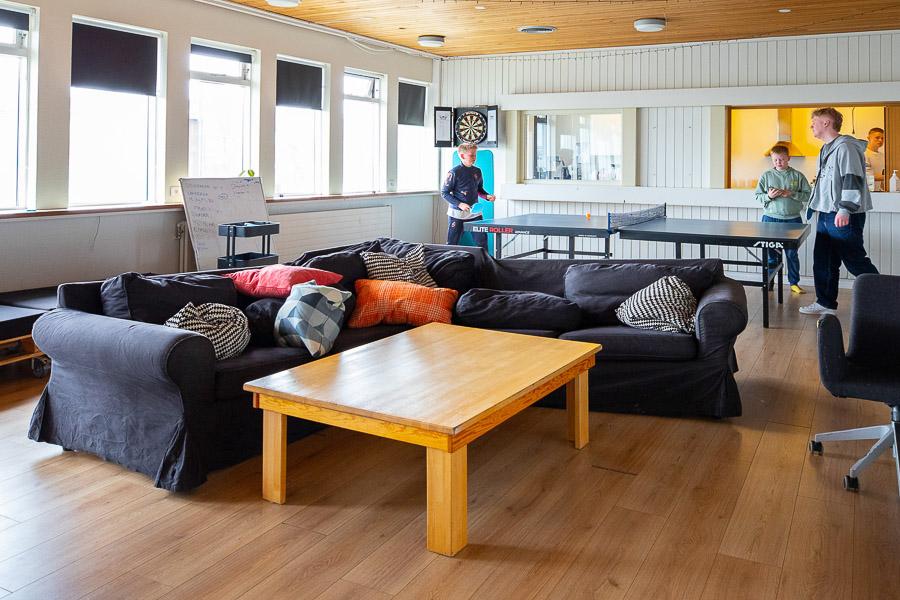 A room with a corner sofa, table and ping-pong table. Teens playing ping-pong. 