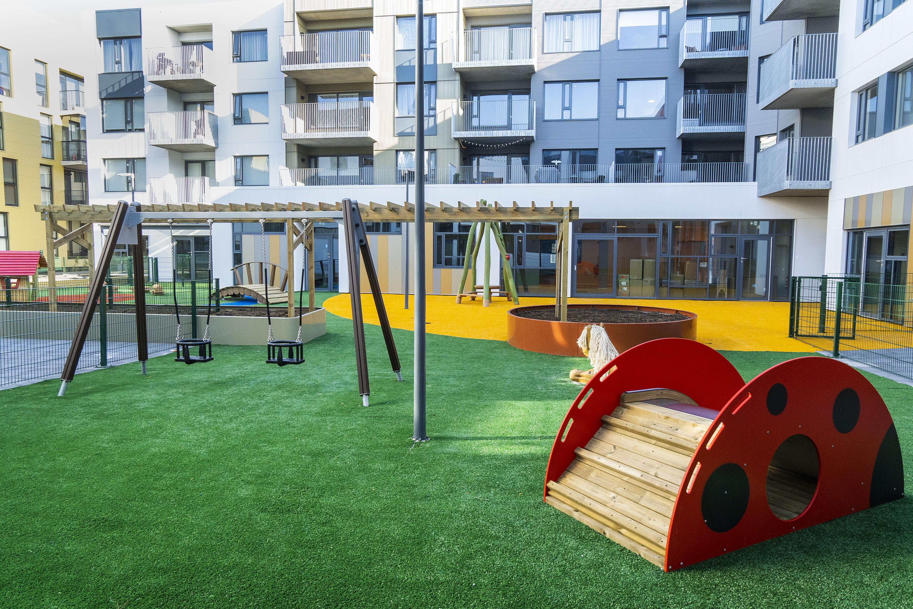 Children's play area with swings and a slide on green and yellow surface. Apartment building housing the Hallgerðargata Nursery School in the background. 