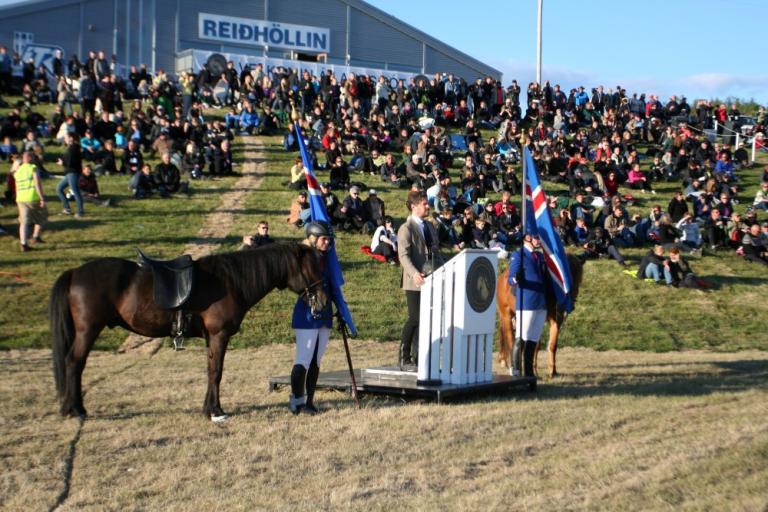 Dagur B. Eggertsson borgarstjóri, á Landsmóti hestamanna í Reykjavík, árið 2012. Fólk situr í brekku, Dagur við ræðupúlt, hestur hjá honum, reiðhöll í bakgrunni.