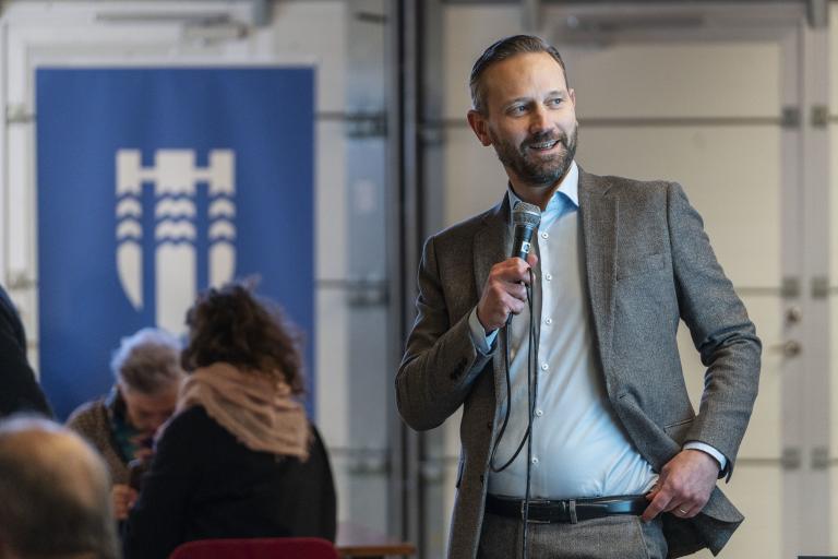 Einar Þorsteinsson borgarstjóri talar í míkrófón á fundi með íbúum. Í bakgrunni er merki borgarinnar.
