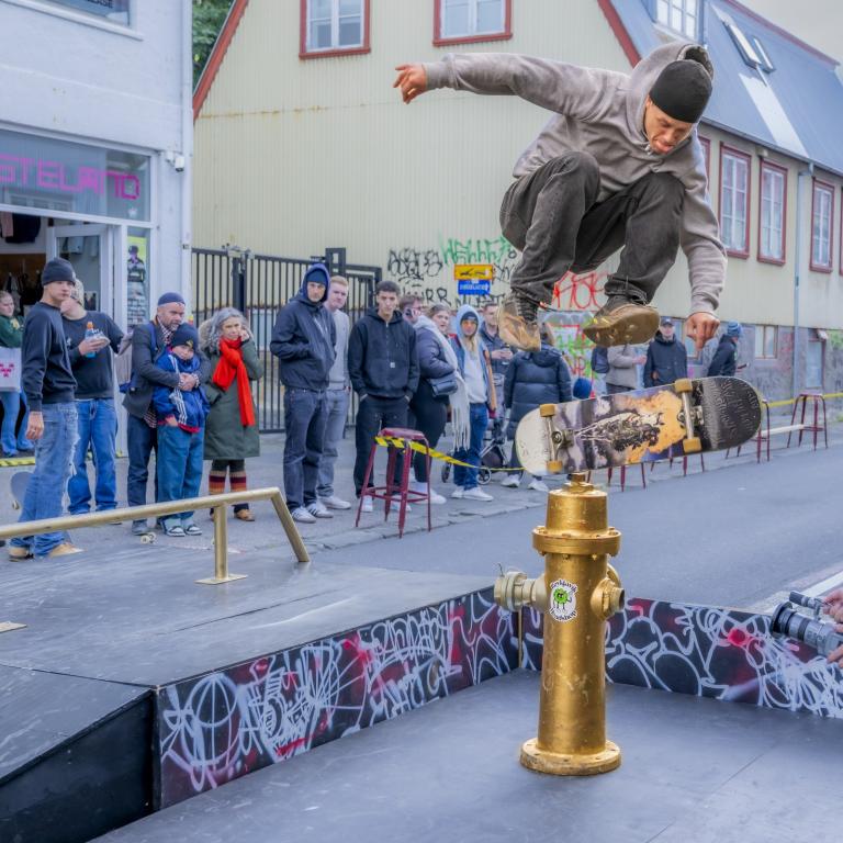 Hjólabrettagaur leikur listir sínar á Hverfisgötu, er að stökkva yfir gylltan vatnshana, fólk horfir í bakgrunni
