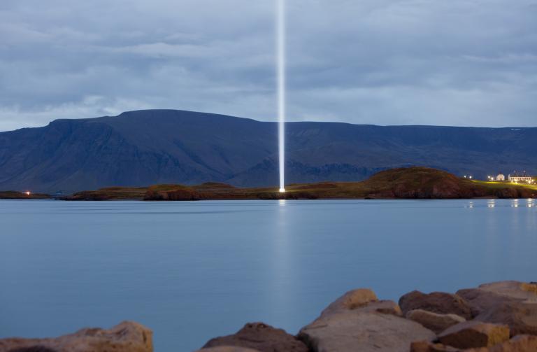 Friðarsúlan í Viðey, séð frá Skarfabakka eða Sæbraut líklega. Skuggsýnt en ekki myrkur, Esjan í baksýn.