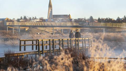 Tvær manneskjur á gangi í Vatnsmýrinni í vetrarstillum. Hallgrímskirkja og göngubrúin yfir Miklubraut í baksýn.