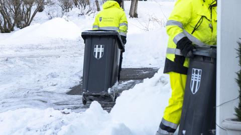 Two waste management employees pull bins marked with the city's logo, with snow outside.
