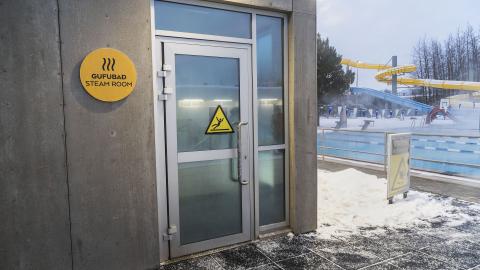 Sauna í Breiðholtslaug, myndin sýnir hana að utan, hurðina inn og skiltið við hlið hurðar. Sundlaugin í bakgrunni. Vetur, snjór.