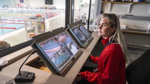 Sundlaugarvörðurinn Dagbjört Ýr Gísladóttir vaktar skjái í vaktherbergi Laugardalslaugar. Situr í rauðri peysu, gluggi út að innilaug vinstra megin í mynd.