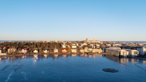Aerial view of Tjörnin and Tjarnargata, seen from Þingholt. The blue waters of Tjörnin are in the foreground, with a blue sky in the background. Calm conditions, clear sky.