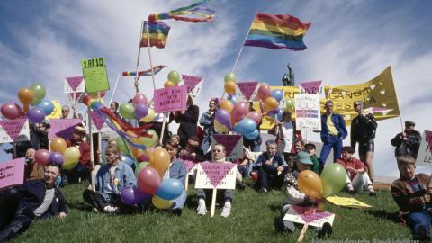 A colorful group of people holding rainbow flags and signs gathers on a grassy area, likely Arnarhóll. Clear weather with a bright blue sky overhead.